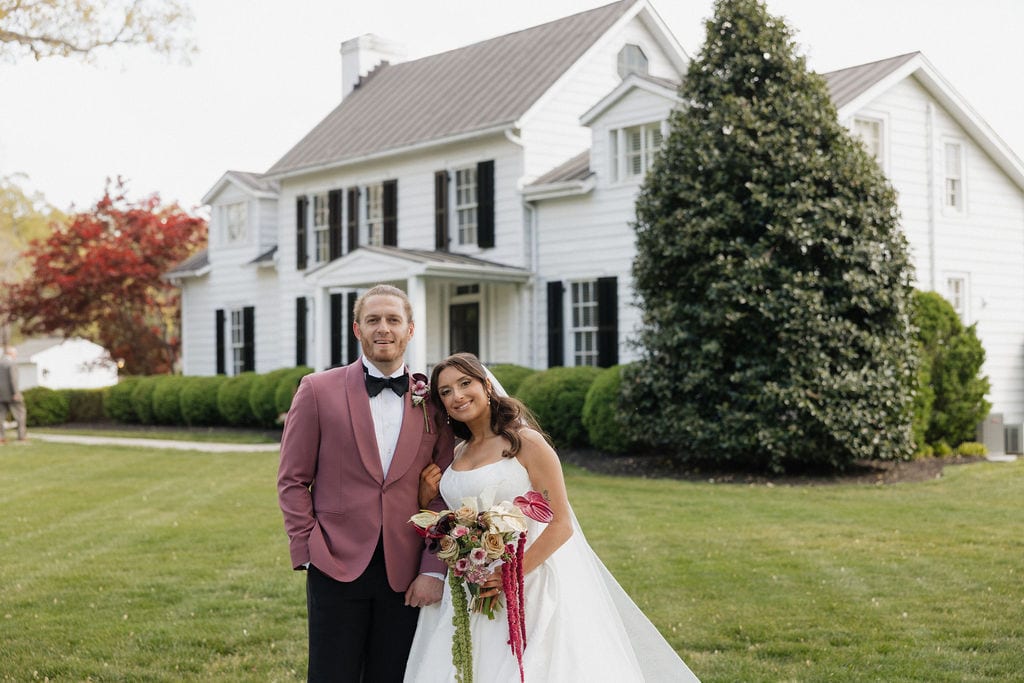 Couple with a rose and anthurium bouquet before the farmhouse at the Callie & Jeremy wedding — designed by Lael Florals