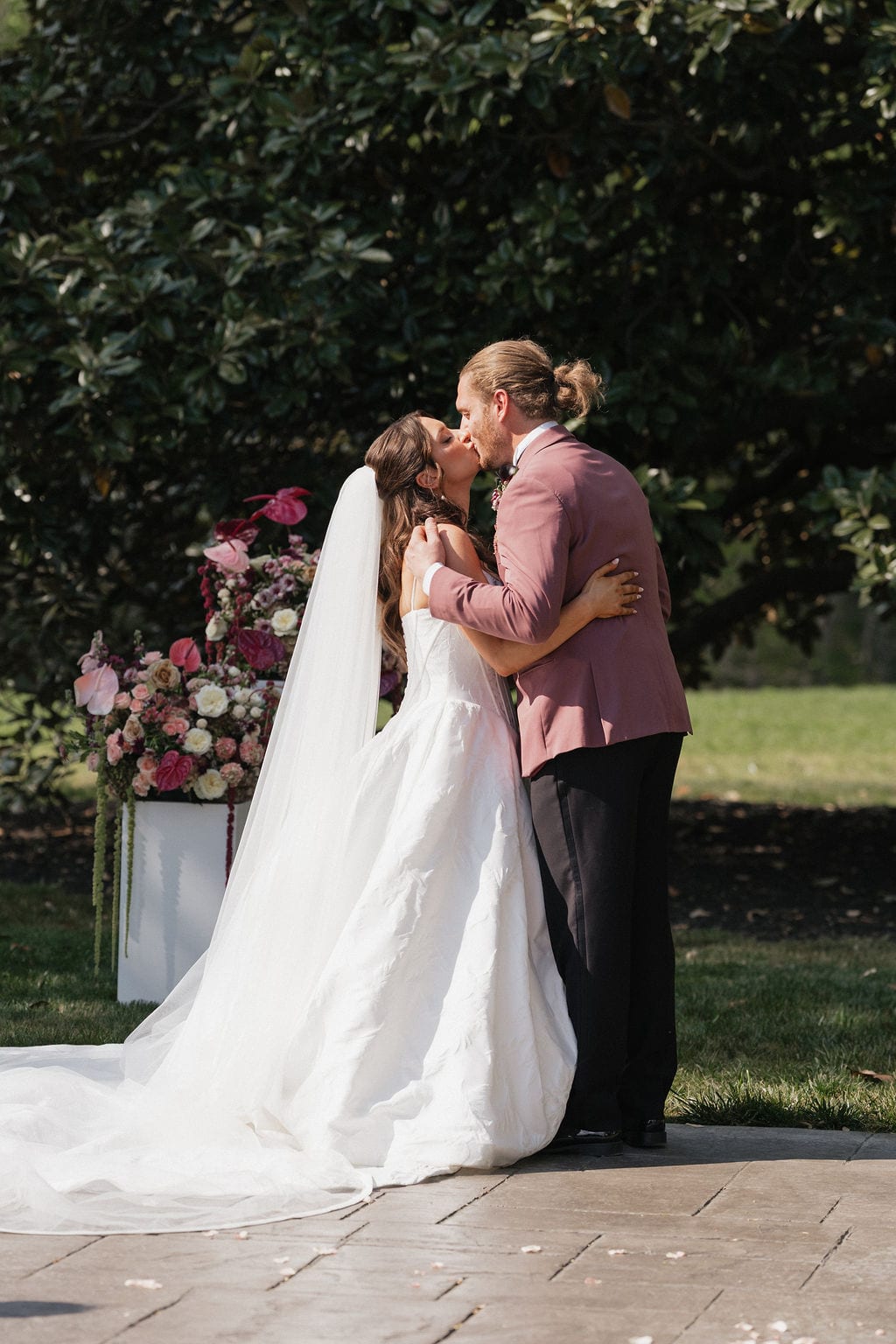 First kiss beside a blush pedestal arrangement at the Callie & Jeremy wedding — designed by Lael Florals