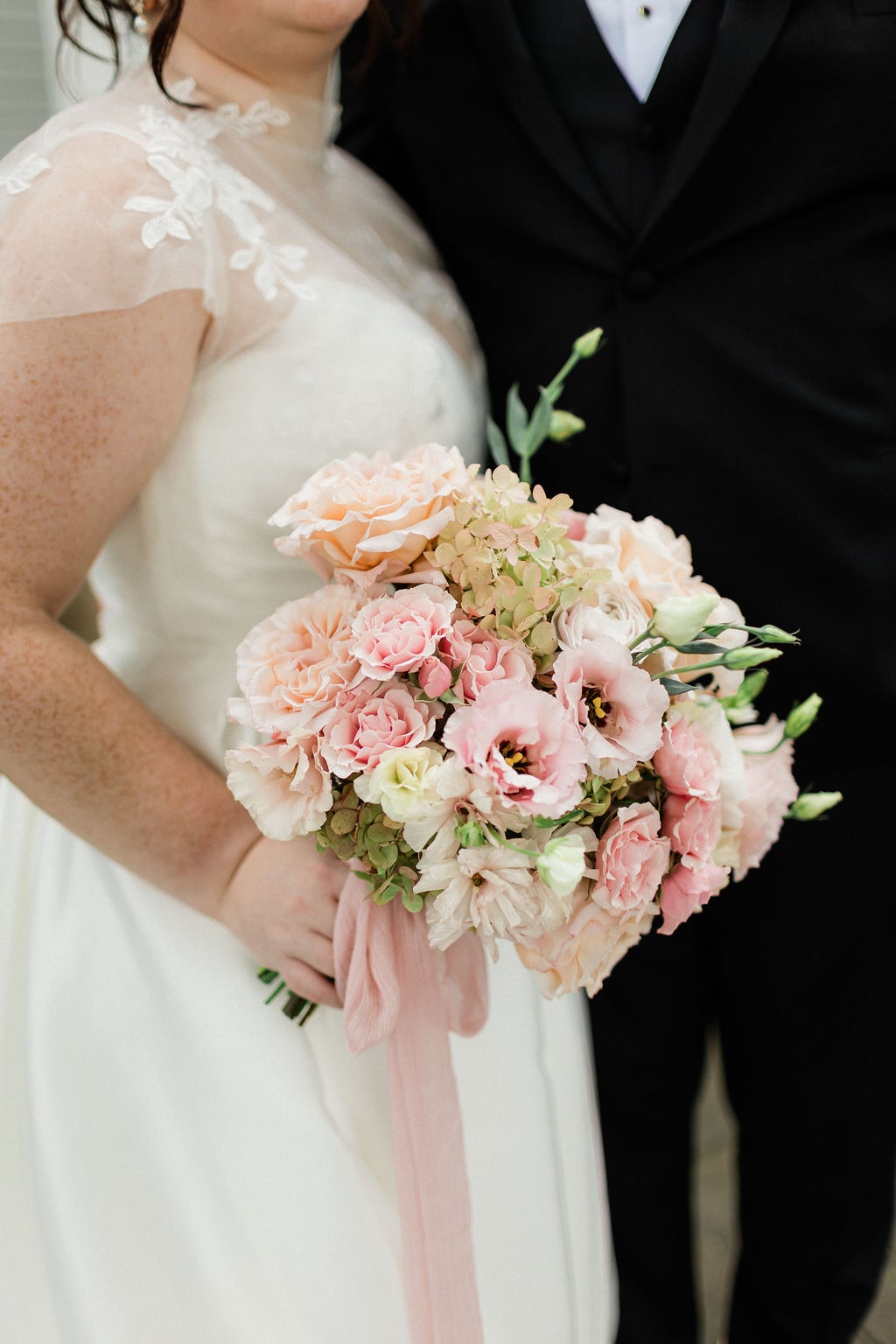The bridal bouquet of blush lisianthus, garden roses and ivory ranunculus with a trailing pink ribbon at the Emily & Thomas wedding — designed by Lael Florals