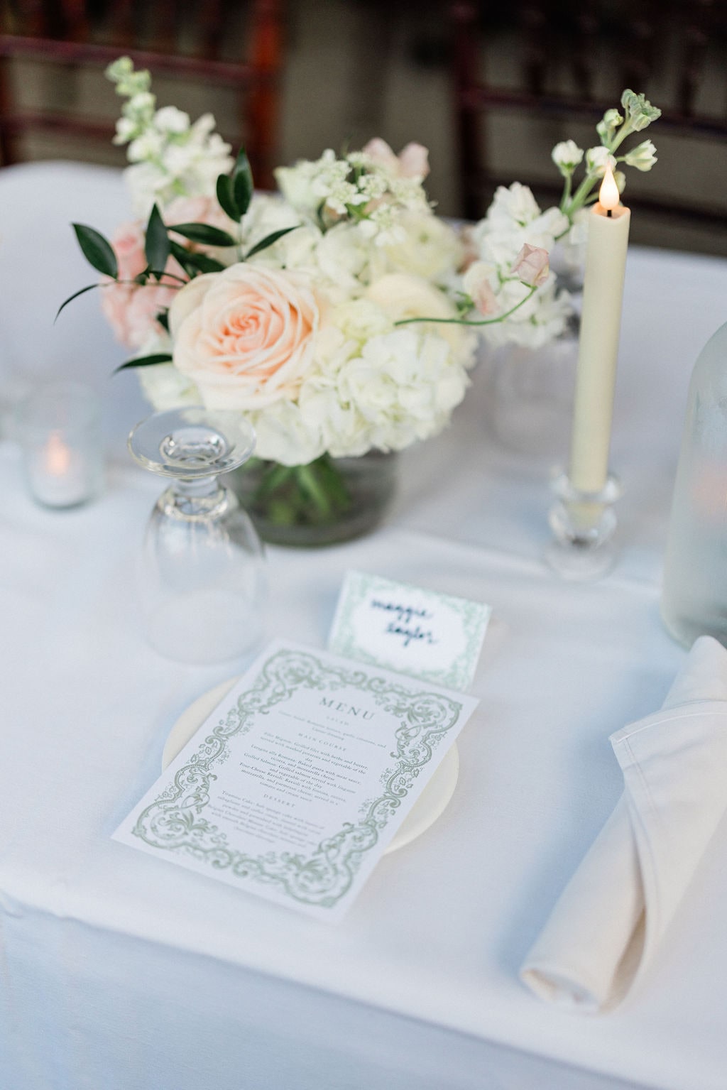 A centerpiece of white hydrangea, blush garden rose and stock beside a taper and menu card at the Emily & Thomas wedding — designed by Lael Florals