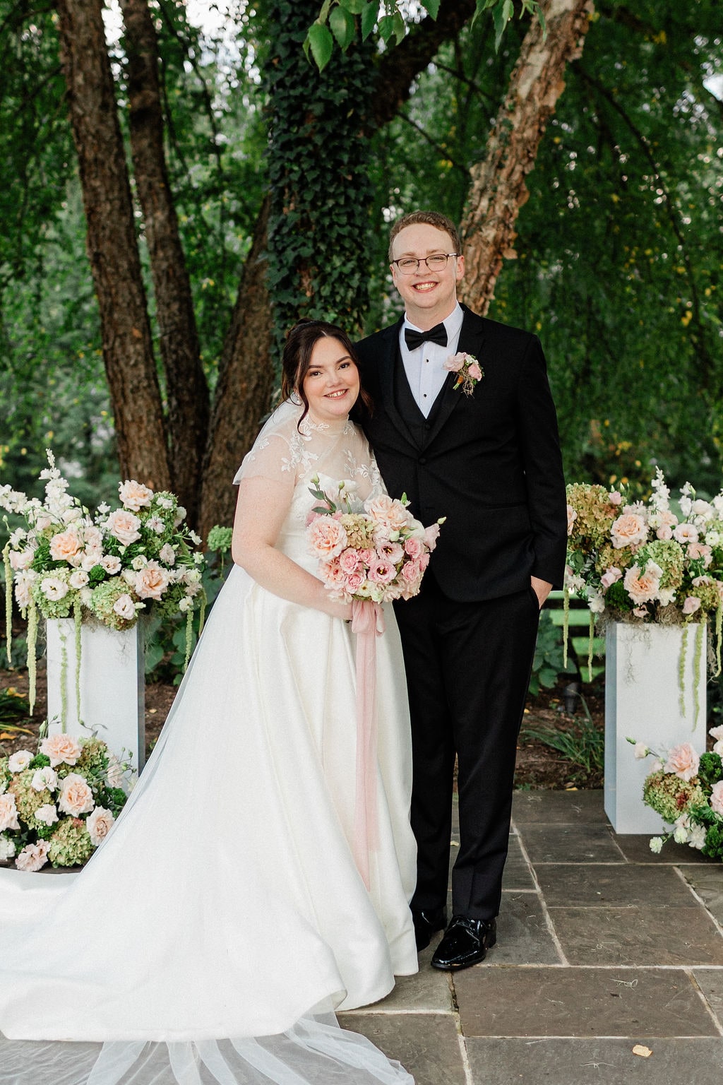 The couple straight on between the pedestal arrangements, the bridal bouquet in hand, at the Emily & Thomas wedding — designed by Lael Florals