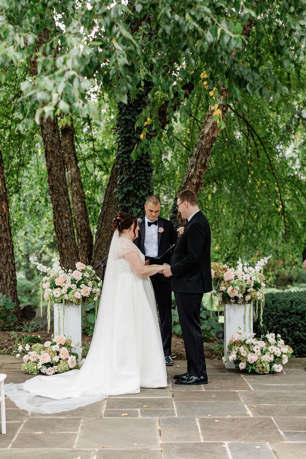 Hands joined at the altar between a symmetrical pair of pedestal arrangements at the Emily & Thomas wedding — designed by Lael Florals