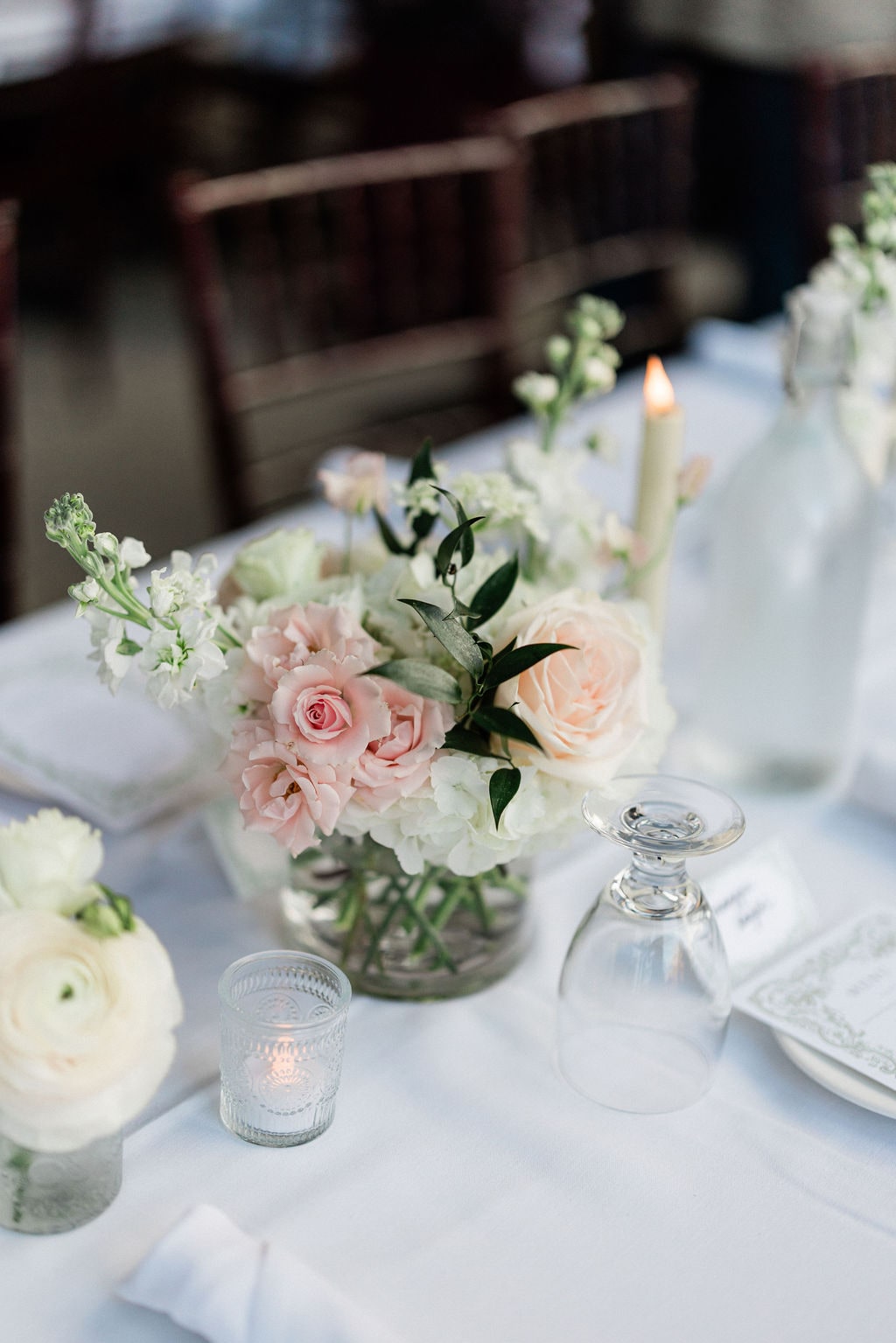 A low arrangement of blush roses and white ranunculus among votives and a taper at the Emily & Thomas wedding — designed by Lael Florals