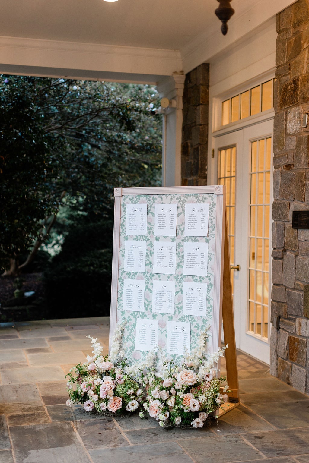 The seating chart easel with a blush and white floral base at its foot at the Emily & Thomas wedding — designed by Lael Florals