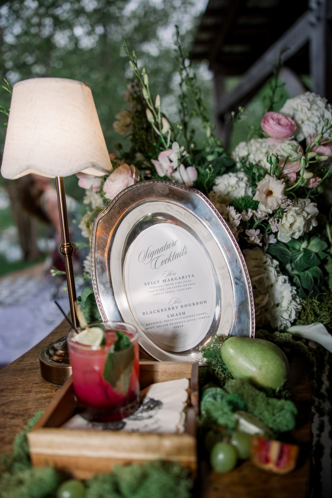 Bar arrangement of ranunculus and hydrangea with fruit framing the cocktail menu at the Gabby & Matt wedding — designed by Lael Florals