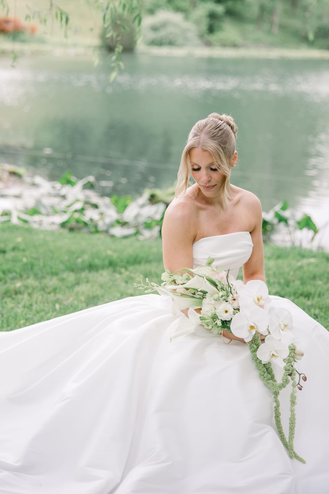 Bride seated lakeside with her cascading orchid bouquet at the Gabby & Matt wedding — designed by Lael Florals
