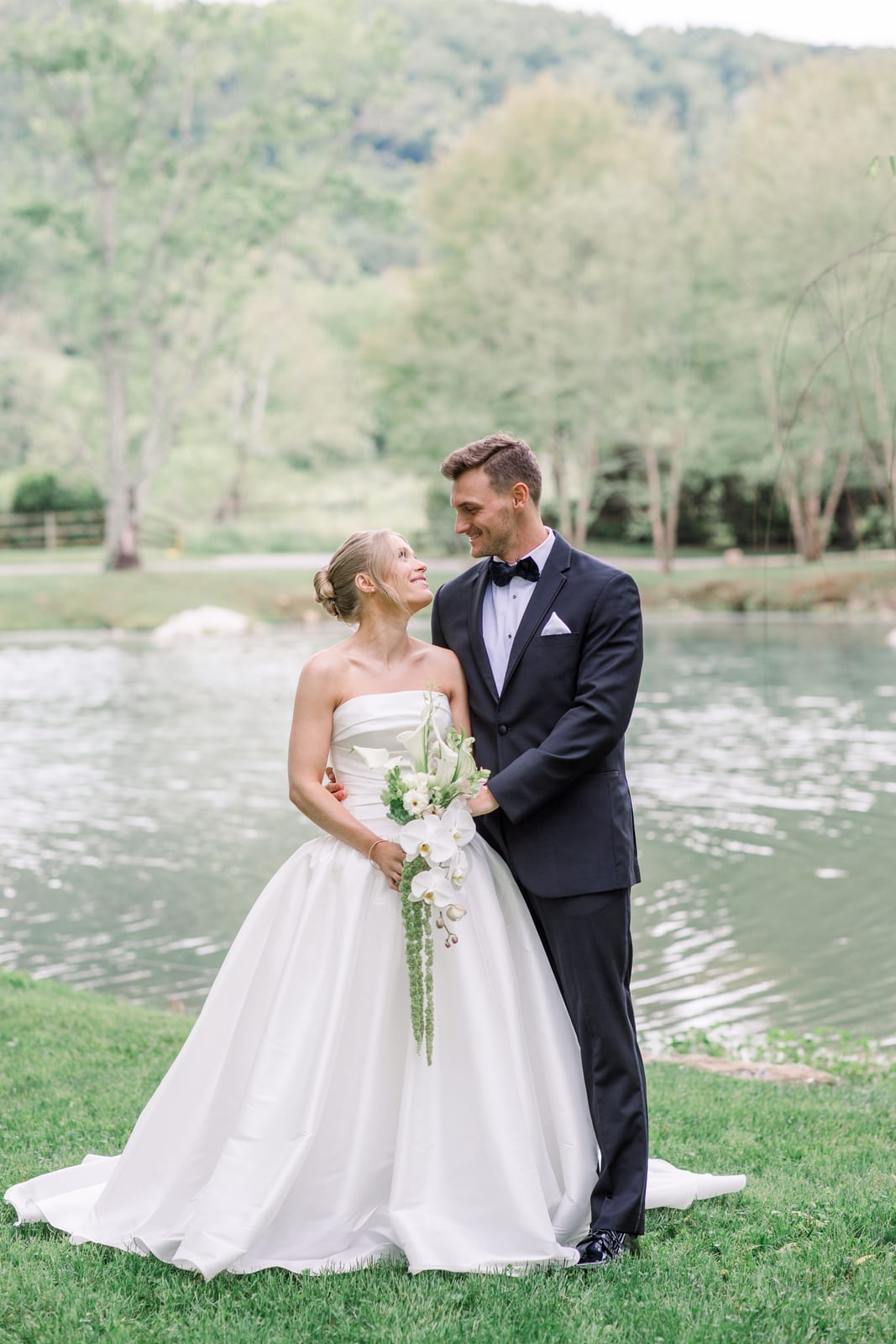 Bride and groom embracing lakeside with a cascading white orchid bouquet at the Gabby & Matt wedding — designed by Lael Florals