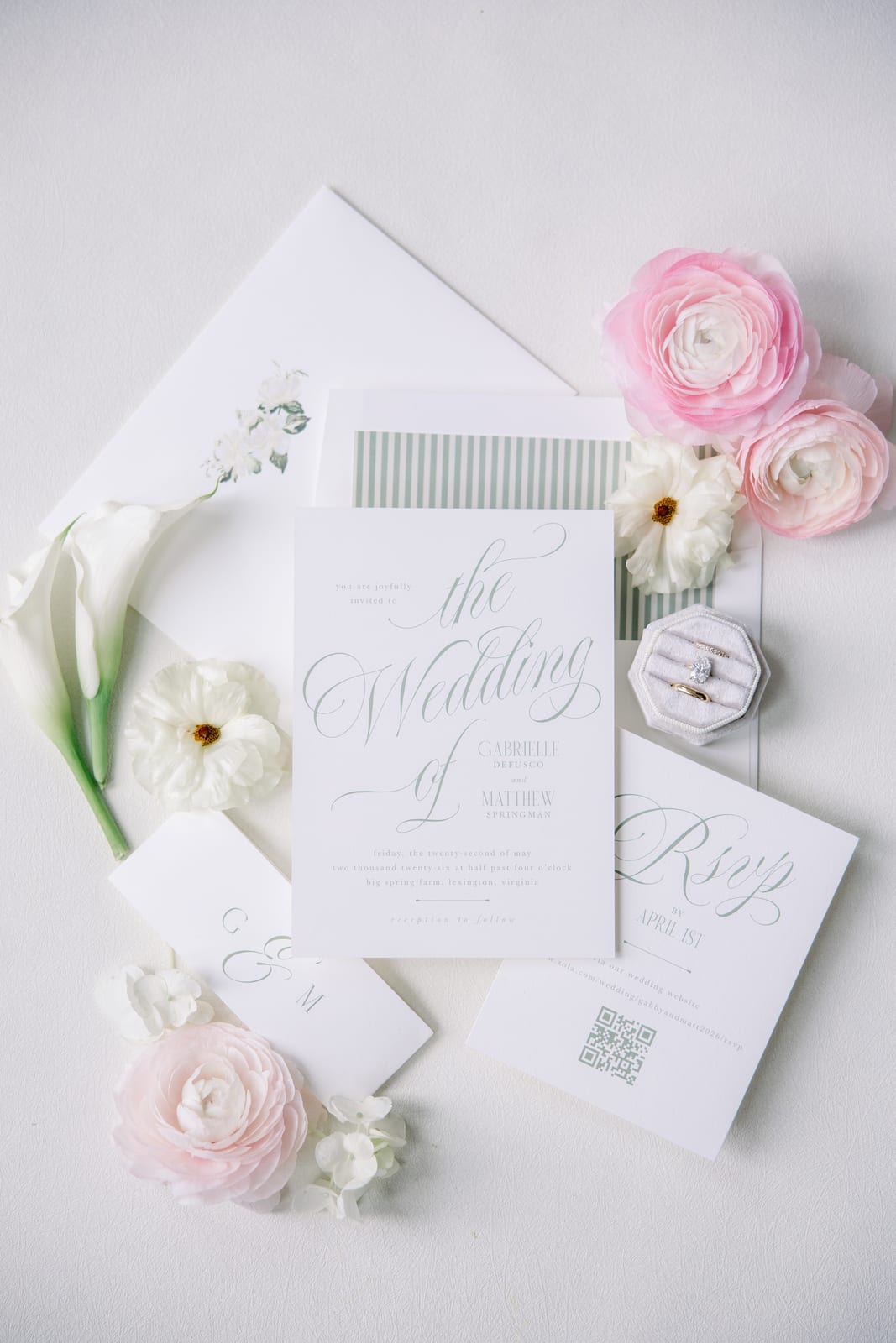 The wedding's real invitation suite styled flat — sage calligraphy on white cards, a striped envelope liner, the velvet ring box, calla lilies and pink ranunculus — from the Gabby & Matt wedding