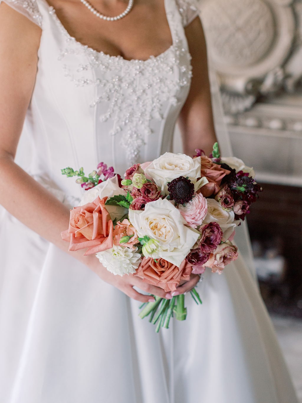 The bridal bouquet of garden roses, burgundy dahlias and cream ranunculus held against the gown at the Karlie & Hank wedding — designed by Lael Florals