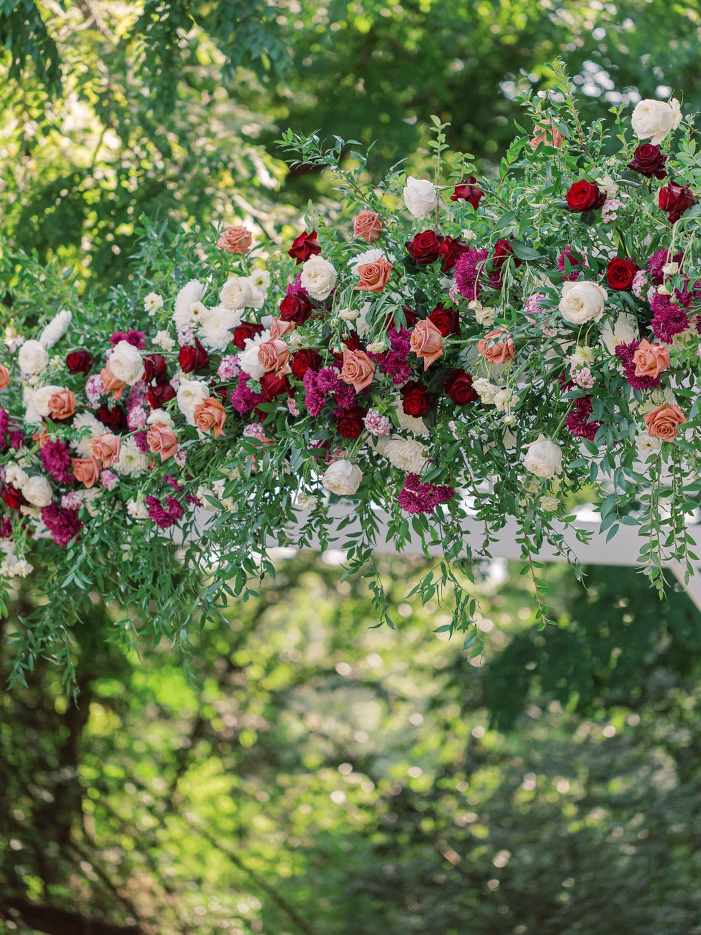 Backlit underside of the pergola garland, roses and burgundy dahlias against open sky, at the Karlie & Hank wedding — designed by Lael Florals