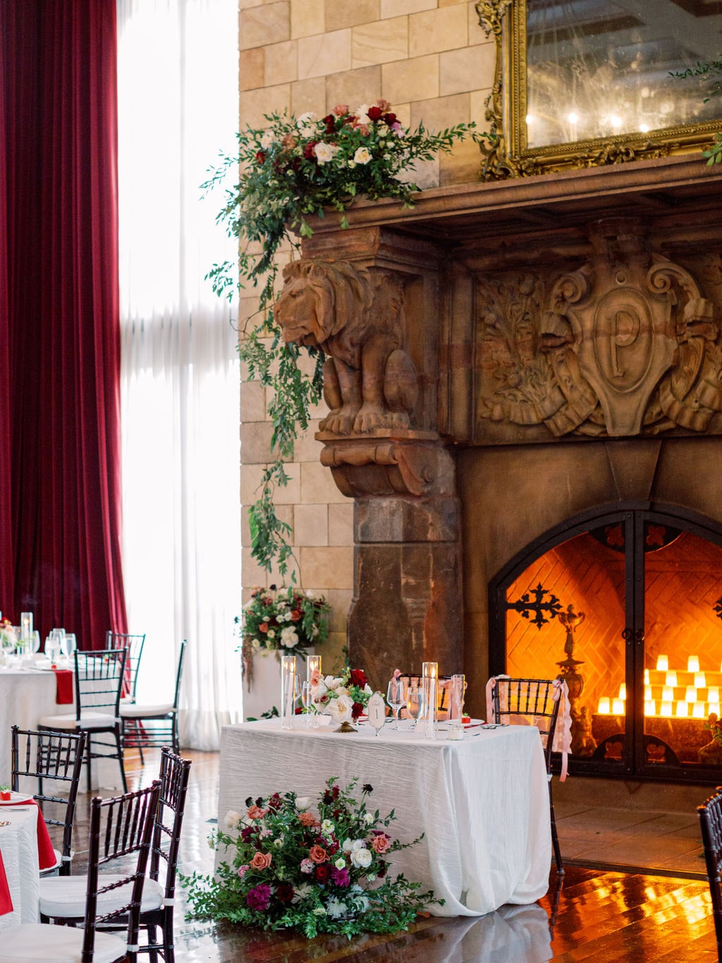 The sweetheart table with a mantel spray above and a full arrangement at its base, fire lit behind, at the Karlie & Hank wedding — designed by Lael Florals