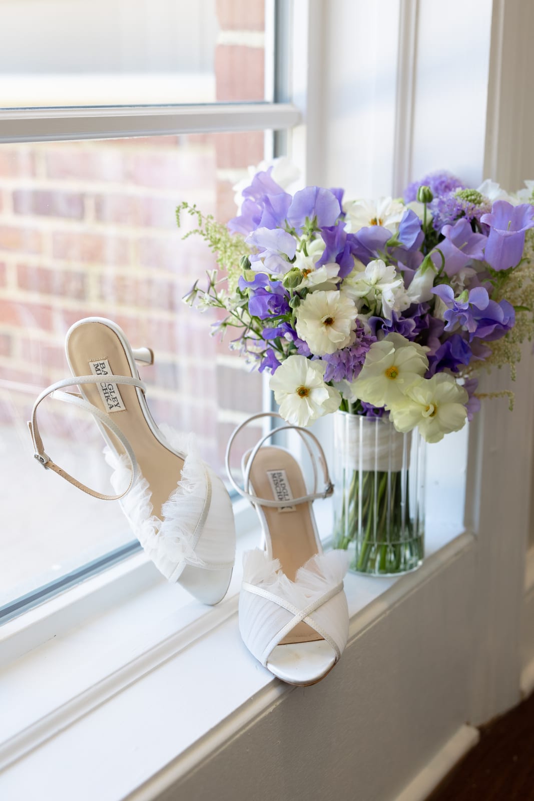 Purple lisianthus gathered loosely in a vase on a windowsill beside the bridal heels — designed by Lael Florals