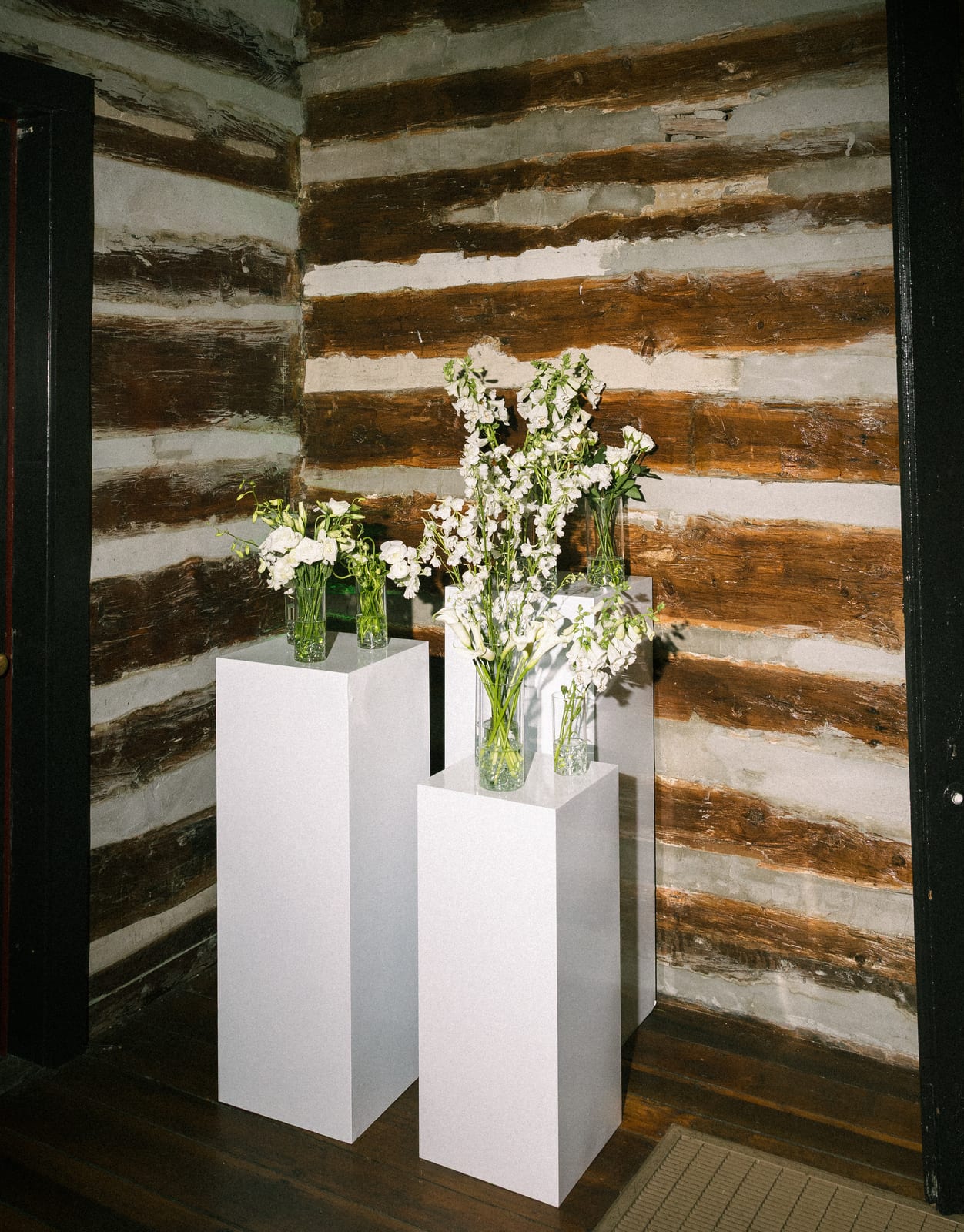 White bud vases on plinths against a log cabin wall at the Sharon & Matt wedding — designed by Lael Florals