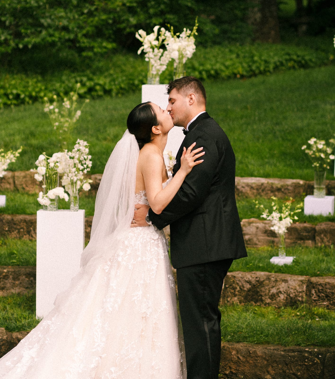 First kiss framed by white floral pedestals on stone steps at the Sharon & Matt wedding — designed by Lael Florals