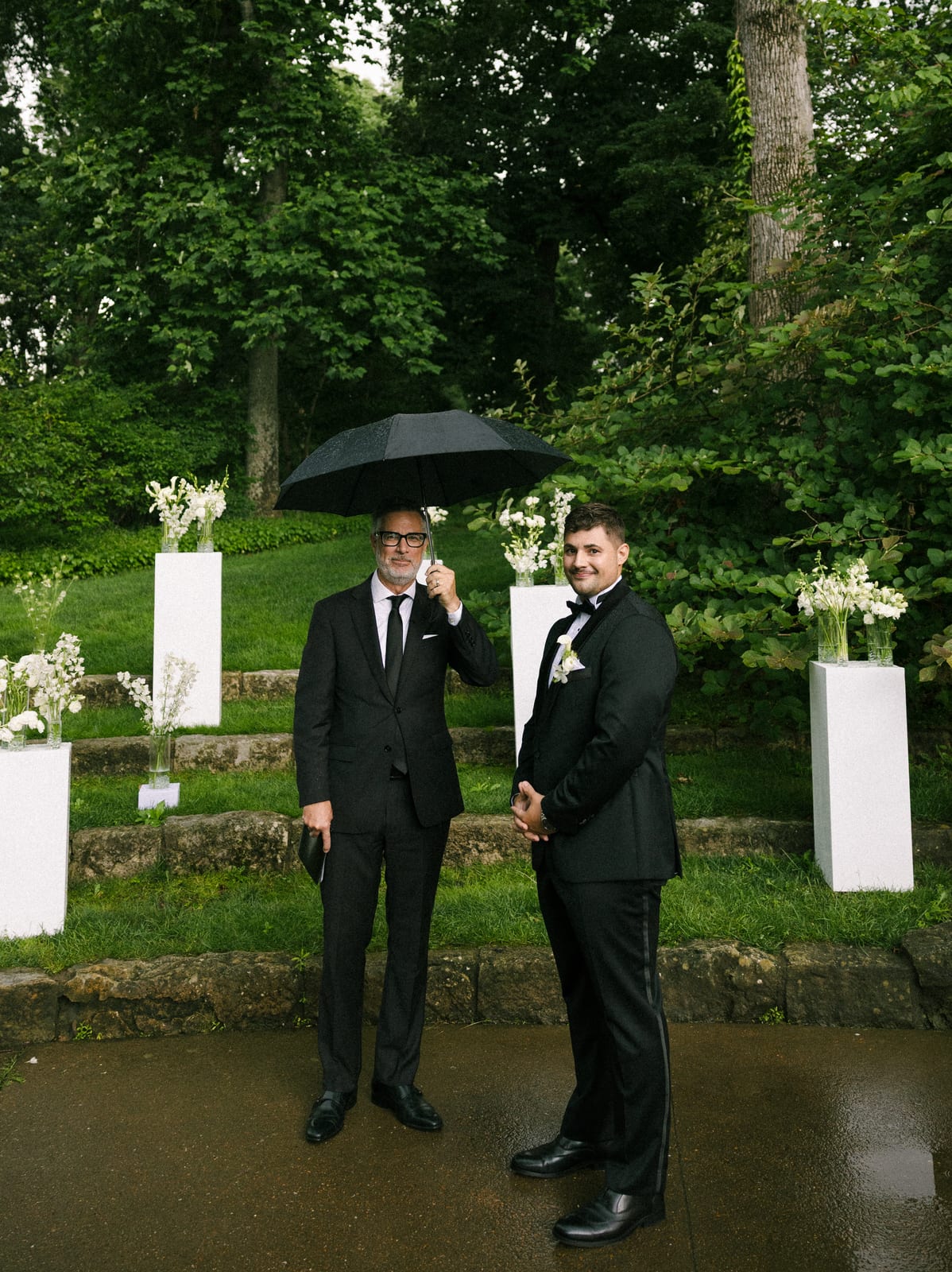 Groom and officiant under an umbrella before pedestal florals at the Sharon & Matt wedding — designed by Lael Florals