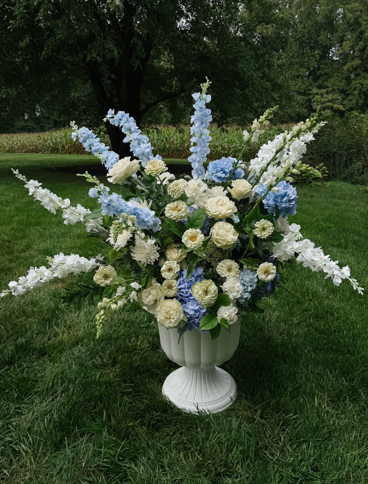 A footed white urn on the lawn, wide and airy with blue delphinium, hydrangea, ivory zinnias, garden roses, and snapdragons