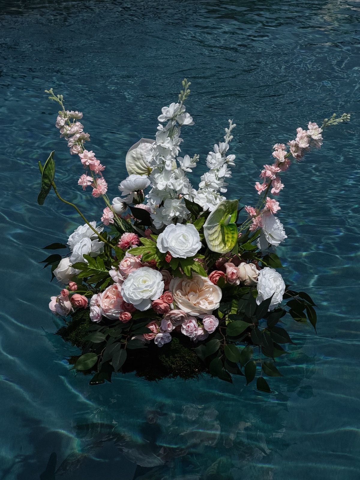 A floating meadow up close: white ranunculus, blush garden roses, anthurium, and pink larkspur spires adrift on the pool