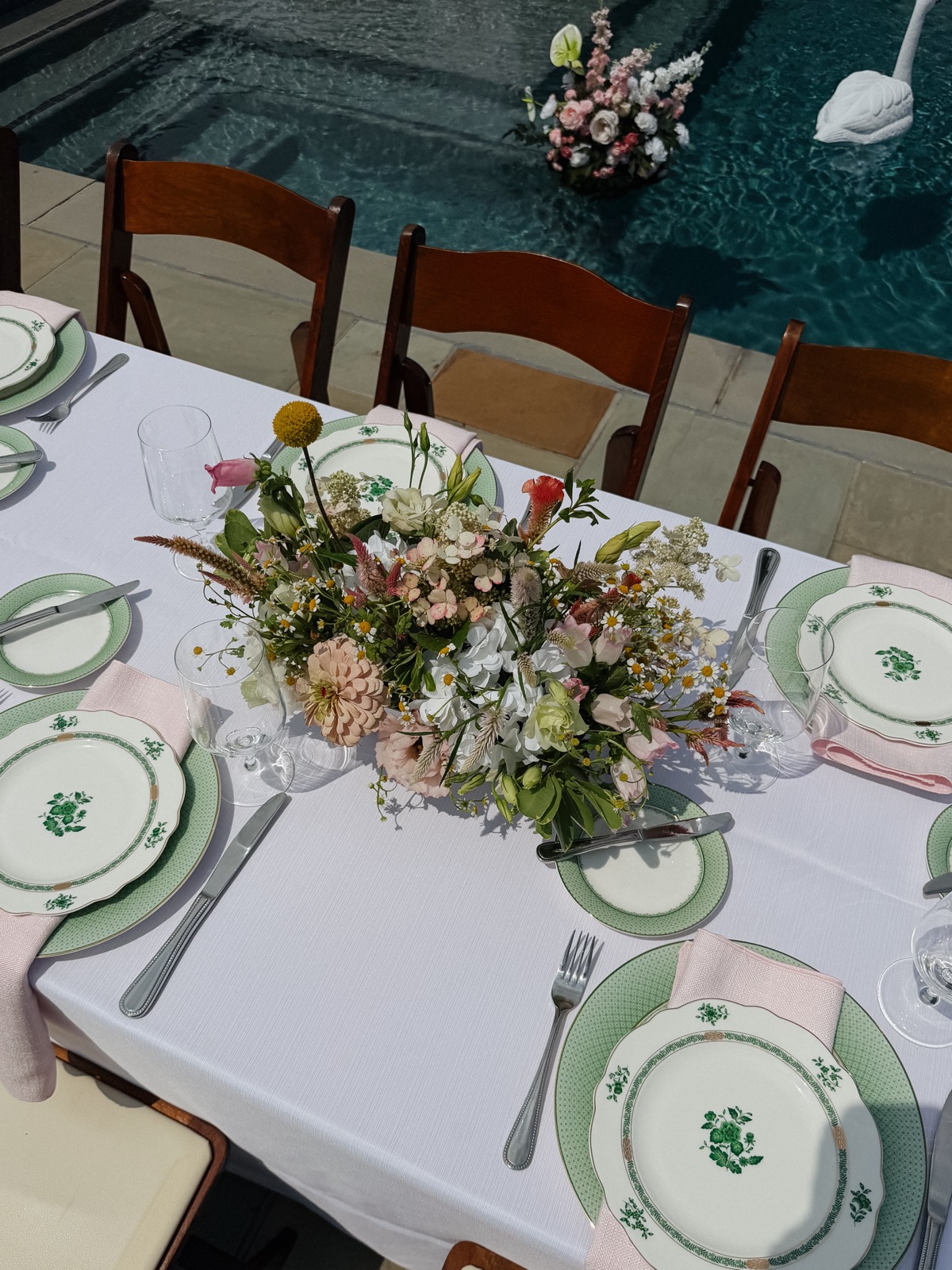 Green-and-white botanical china and pale pink napkins on white linen, a low loose centerpiece of zinnias, chamomile, and hydrangea, and a floating arrangement drifting in the pool beyond