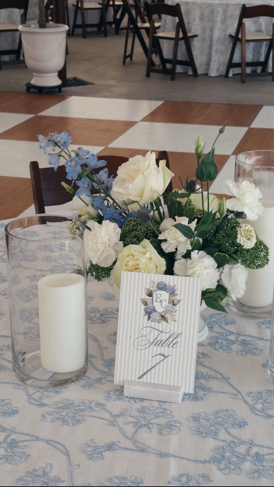 A compact centerpiece of blue delphinium, cream roses, and carnations on blue-and-white embroidered linen, flanked by pillar candles in glass hurricanes beside the monogrammed Table 7 card, the checkerboard dance floor beyond
