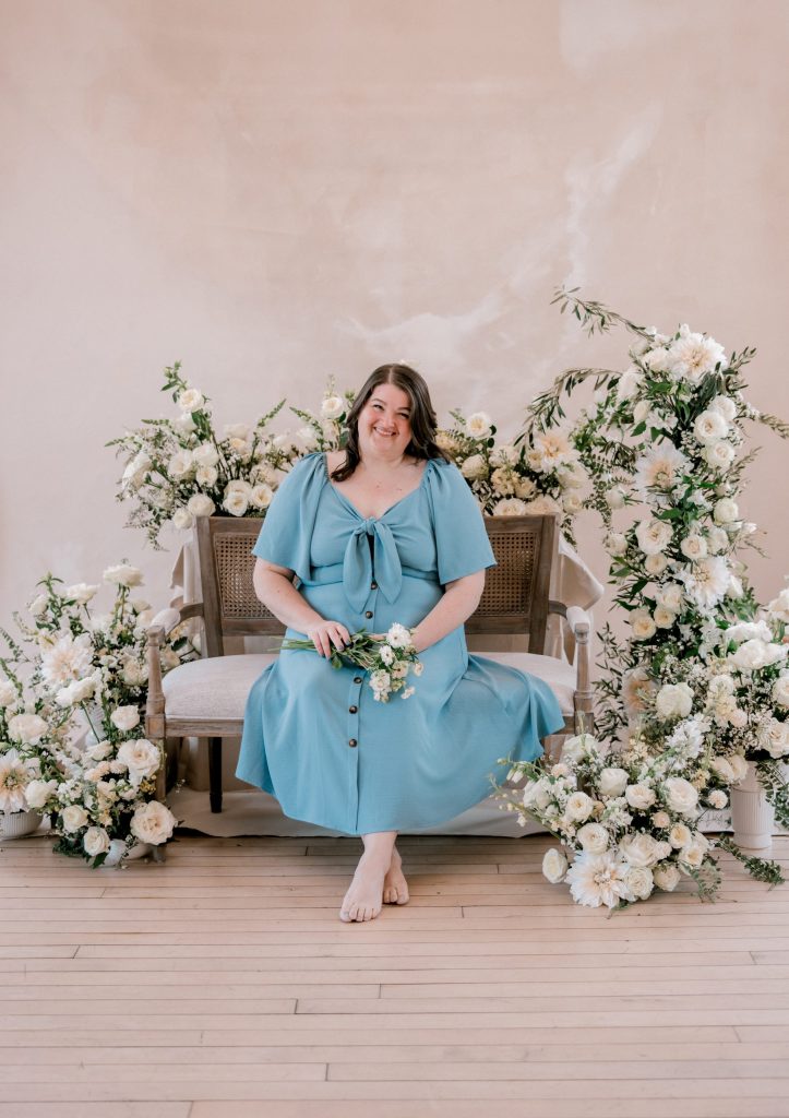 Jamie of Lael Florals, smiling in a blue dress, sits on a rustic bench framed by large, romantic floral arrangements of white flowers and greenery.