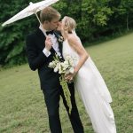 A groom in a black tuxedo holds a white paper parasol over his bride as they kiss in a grassy field. The bride holds a bouquet of white flowers and long, trailing greenery.