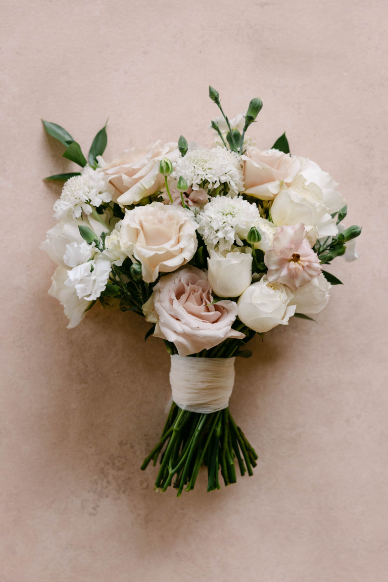 A romantic wedding bouquet, seen from directly above, rests on a pale tan surface. It is filled with blush pink, pale peach, and ivory roses, white scabiosa (pincushion flowers), and delicate white lisianthus, all interspersed with dark green leaves. The gathered green stems are wrapped tightly in an ivory fabric ribbon.