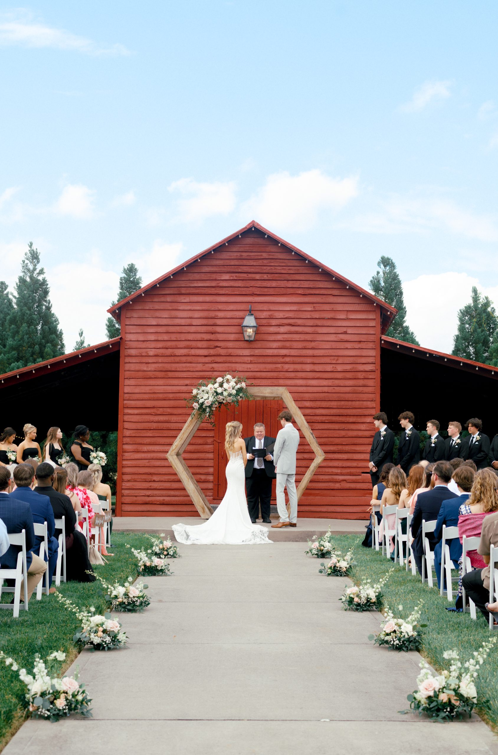 Alt text: An outdoor wedding ceremony in front of a red barn with a wooden hexagon arch adorned with blush and white flowers. The bride in a white gown and the groom in a light gray suit stand at the altar, surrounded by bridesmaids in black dresses and groomsmen in black suits. Guests are seated in white chairs along an aisle lined with small floral arrangements.