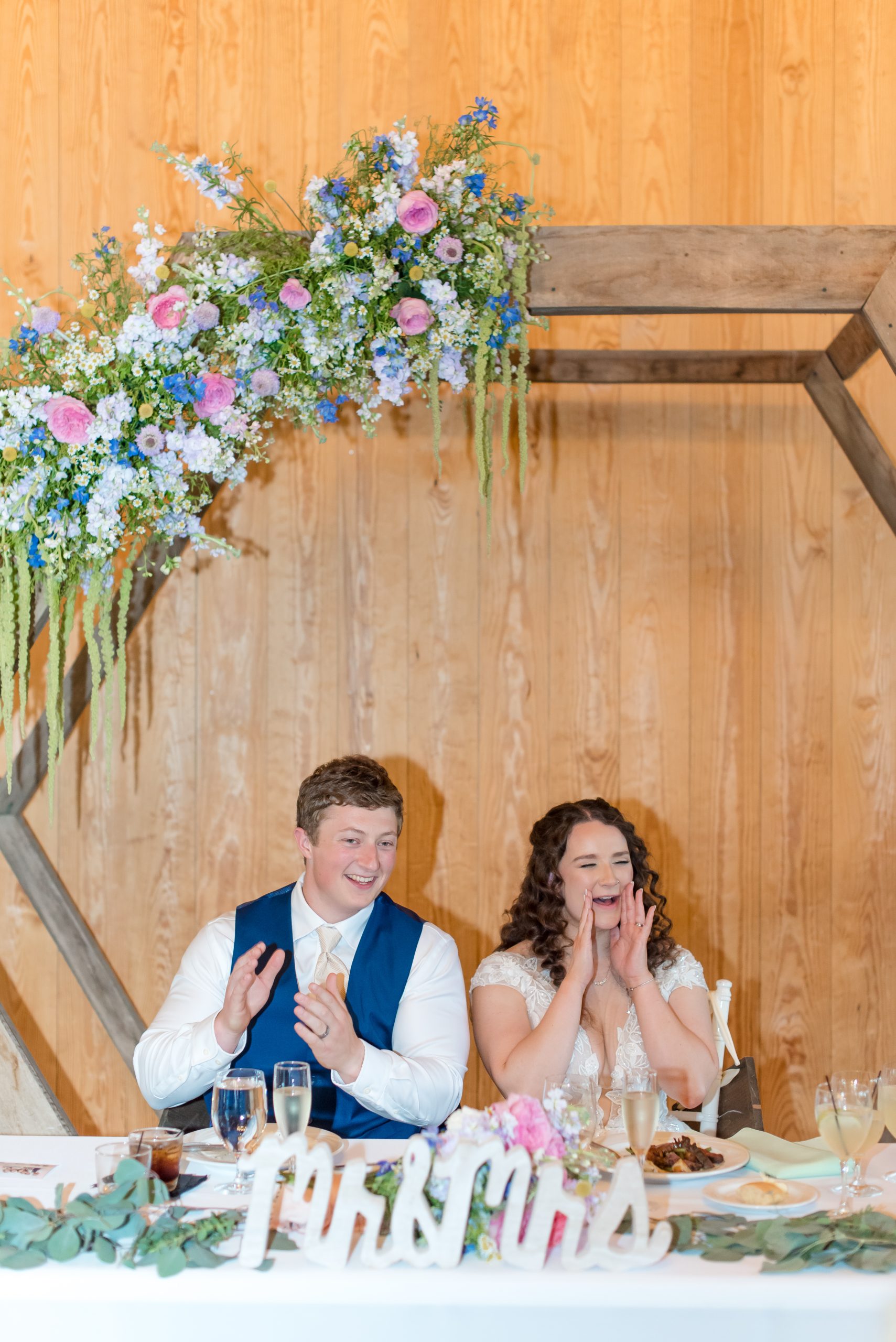 The bride and groom sit at their sweetheart table inside The Barns of Kanak, smiling and clapping during the reception. Behind them, a wooden hexagon arch is adorned with cascading pastel florals in pink, blue, and white. The table is decorated with greenery and a “Mr & Mrs” sign.