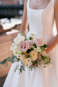A close-up shot of a bride wearing a modern square-neck wedding gown holding a lush bouquet. The floral arrangement features white garden roses, dusty mauve roses, and white ranunculus mixed with textured greenery.