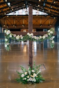 A large, dark-stained wooden cross standing on a polished floor in an industrial-style venue. The crossbar is draped with a lush garland of greenery, white roses, and dusty mauve roses that hang down at the ends. A matching floral arrangement sits at the base of the cross. The background features high ceilings with exposed metal trusses and blurred architectural details.