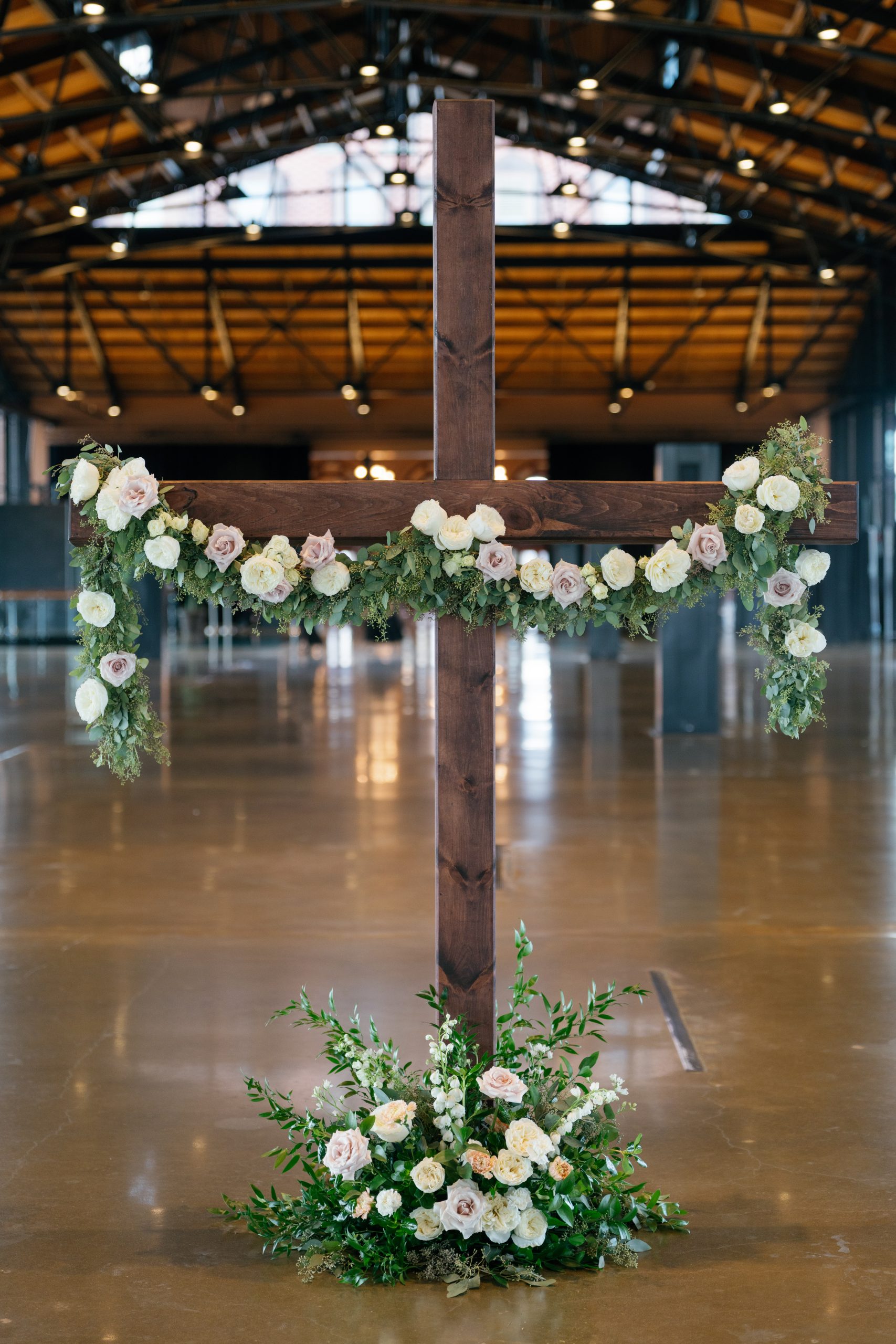 A large, dark-stained wooden cross standing on a polished floor in an industrial-style venue. The crossbar is draped with a lush garland of greenery, white roses, and dusty mauve roses that hang down at the ends. A matching floral arrangement sits at the base of the cross. The background features high ceilings with exposed metal trusses and blurred architectural details.