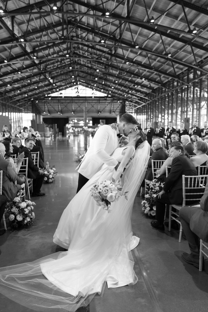 A black-and-white photograph of a groom in a white tuxedo jacket dipping his bride for a romantic kiss in the center of the wedding aisle. The bride, holding a bouquet, leans back while guests seated on either side clap and smile. The background features a large industrial venue with exposed ceiling trusses and a wooden cross in the distance.