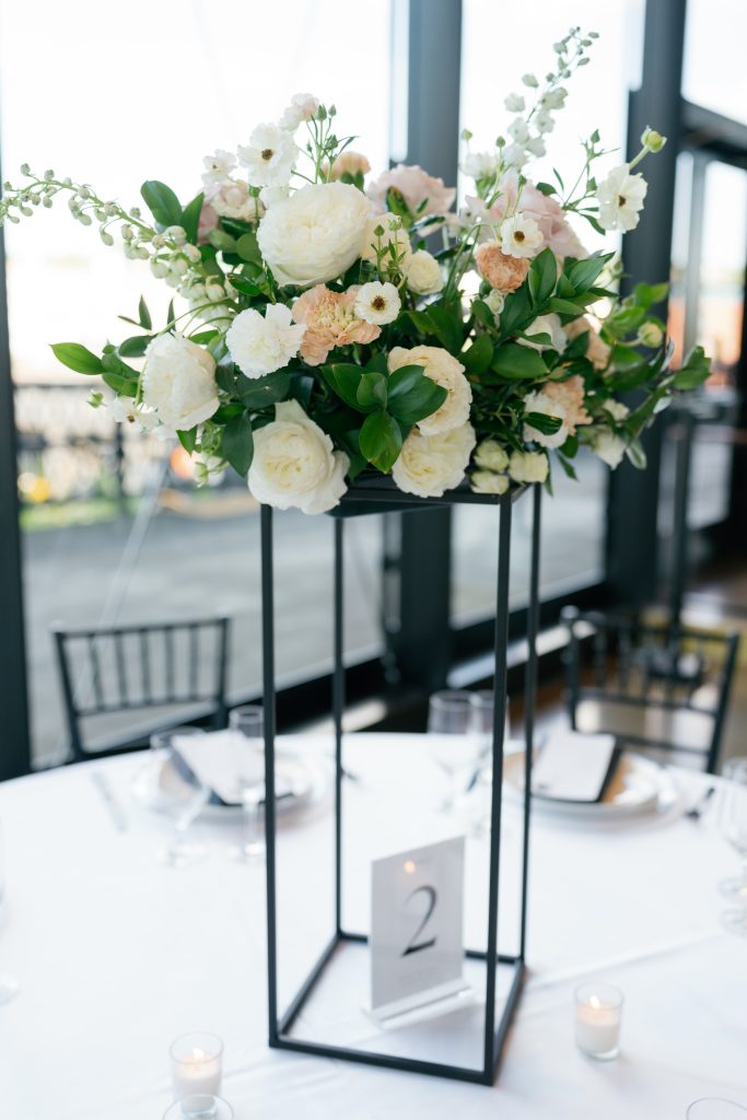 A tall wedding centerpiece features white roses, peach carnations, and greenery atop a black metal geometric stand on a round table. The table setting includes a white tablecloth, black napkins, votive candles, and a table number "2" card. Large industrial windows with black frames are visible in the background.