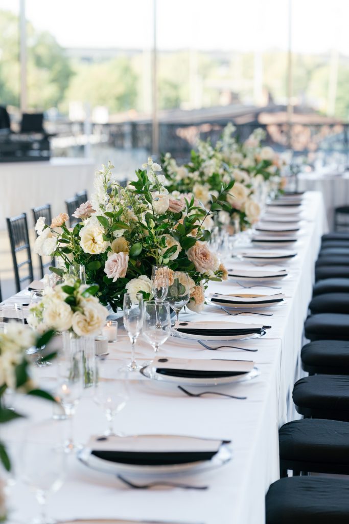 A perspective shot looking down the length of a long wedding reception table set with white linens and black napkins. A continuous floral runner features white garden roses, dusty mauve blooms, and peach carnations mixed with greenery. Black Chiavari chairs line the table, and large industrial windows provide a bright background.