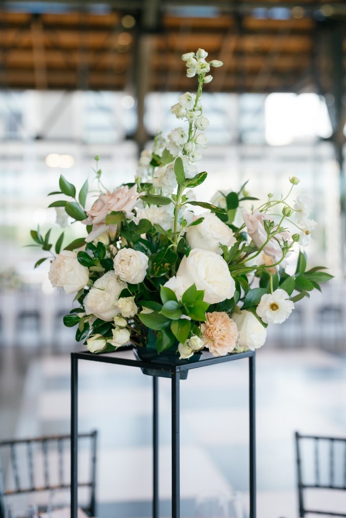 A vertical close-up of a tall wedding centerpiece sitting on a thin black metal geometric stand. The floral arrangement features white garden roses, dusty mauve roses, peach carnations, and tall sprigs of white stock or delphinium reaching upward. The background shows a blurred industrial venue with large windows and high ceilings.