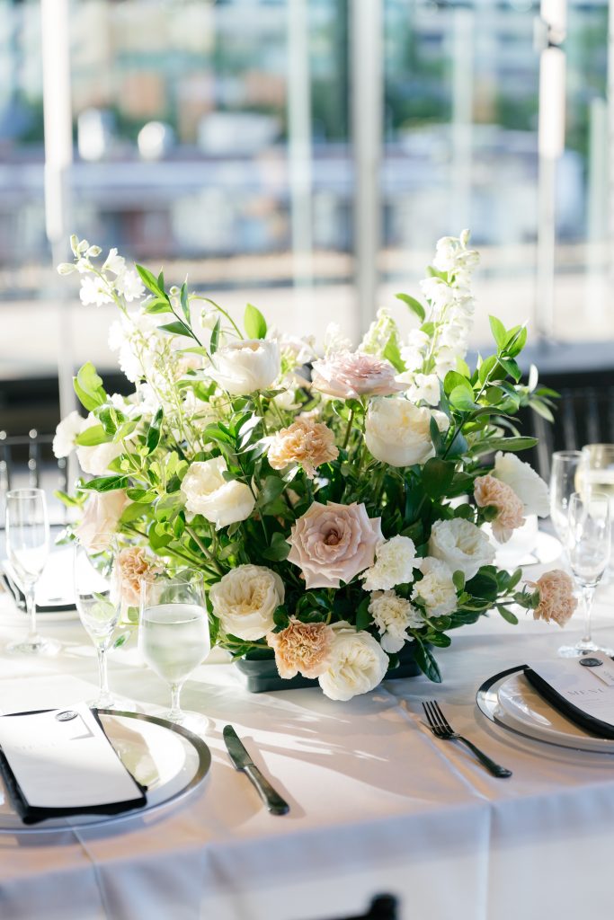 A close-up of a lush, low wedding centerpiece featuring white garden roses, dusty mauve roses, and peach carnations on a white tablecloth. The table setting includes elegant menu cards with dark wax seals, silverware, and crystal stemware catching the bright natural sunlight. Blurred floor-to-ceiling windows are visible in the background.